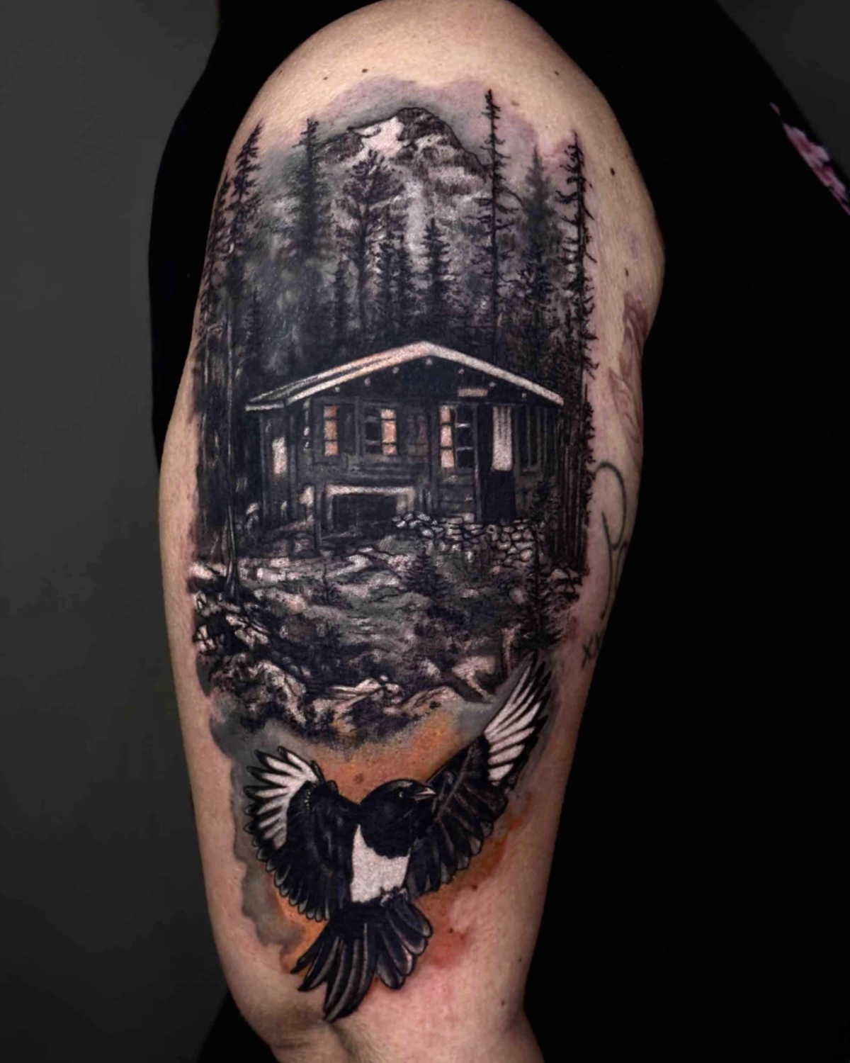 Black & Grey Tattoos Deggendorf — realistische Berghütte im Wald mit fliegender Elster am Oberarm, Tätowierer Deggendorf Luca Limito, Atelier Voltaire Tattoo Studio / Black and grey tattoos Deggendorf — realistic mountain cabin in forest with flying magpie on upper arm by Luca Limito at Atelier Voltaire Tattoo Studio